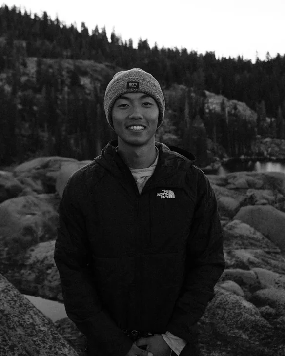 Stephen Hu smiling in a beanie and dark jacket, with granite boulders, a lake and pine trees behind him.